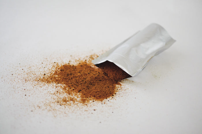 En öppen Mylarpåse med kryddpulver som hälls ut, omgiven av en del av innehållet som sprids ut på en vit yta.